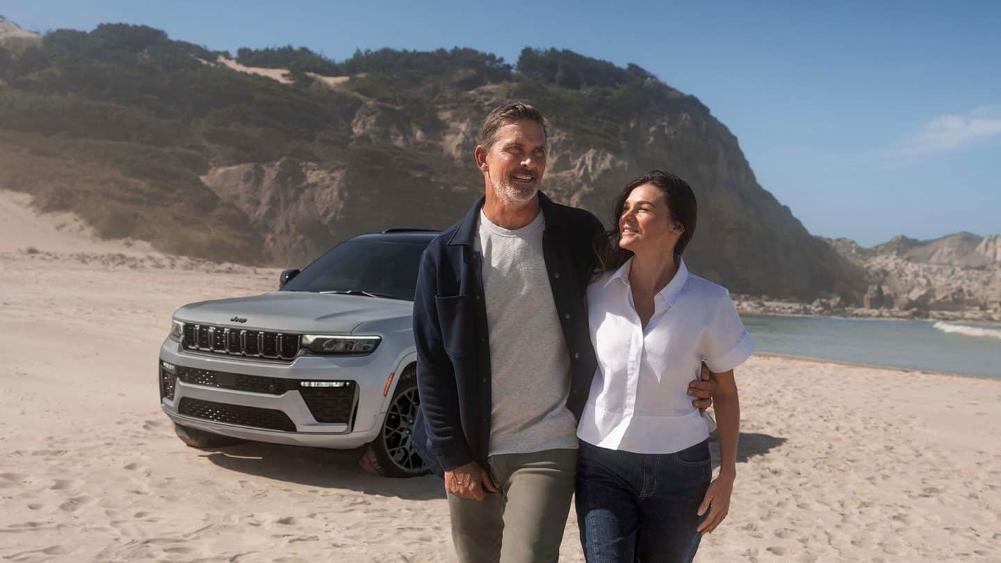 A man and woman walking away from a gray 2027 Jeep Grand Cherokee Summit Obsidian, parked on a sandy beach.
