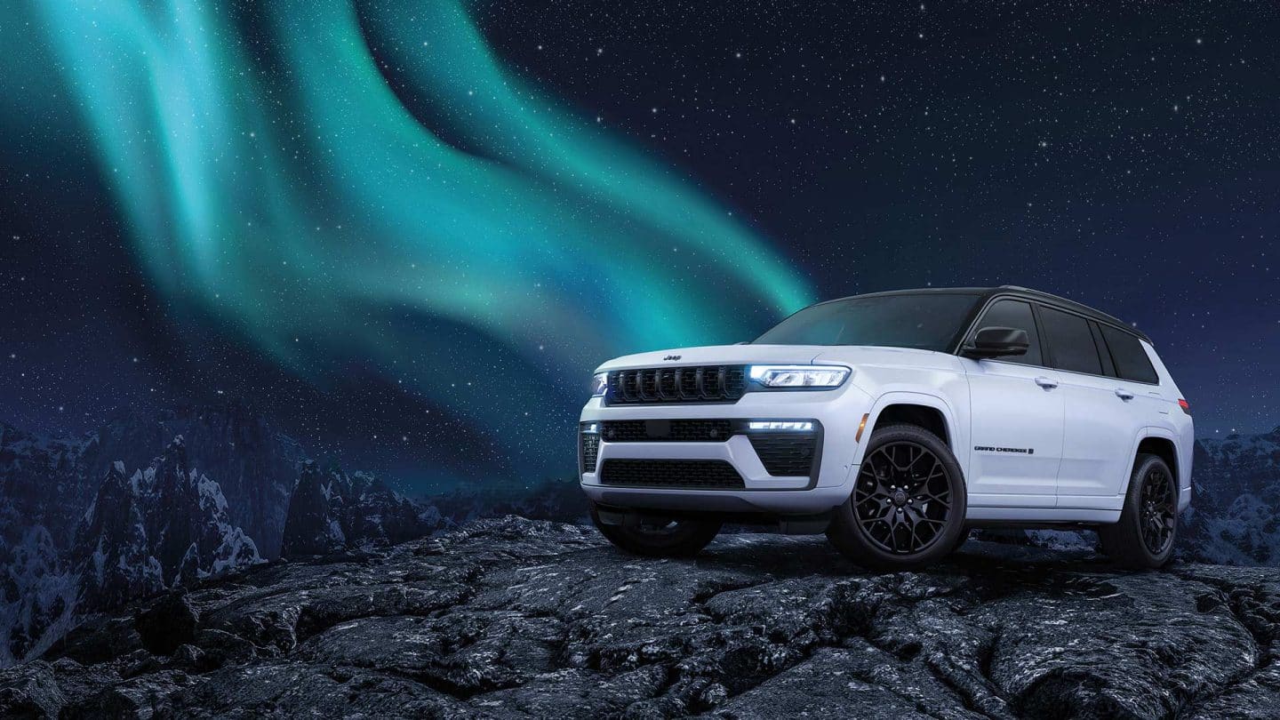 A white and black 2027 Jeep Grand Cherokee Summit Obsidian, parked on a rocky hill at night, with stars and a trail of the northern lights in the background.