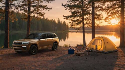 A copper and black 2027 Jeep Grand Cherokee Summit Obsidian parked at a campsite beside a lake at sunset.