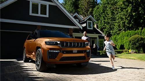 A copper and black 2027 Jeep Grand Cherokee Summit Obsidian parked in the driveway of a residential home with a man and his daughter nearby.
