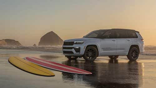 A silver and black 2027 Jeep Grand Cherokee Summit Obsidian parked on a foggy beach at sunrise with two boogie boards nearby.