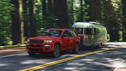 A red 2027 Jeep Grand Cherokee Limited Reserve traveling down a highway in the woods, towing a travel trailer.