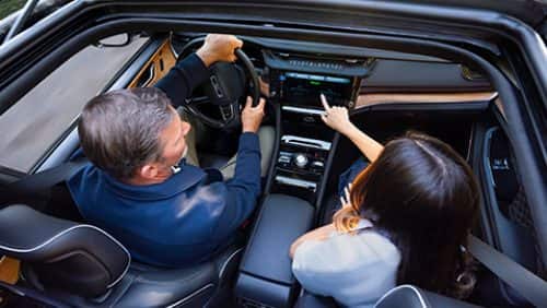 The front seat area of the 2027 Jeep Grand Cherokee Summit, with a man in the driver seat and a woman in the passenger seat. Shot from outside the open sunroof.