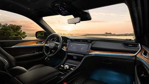 The front seat area of the 2027 Jeep Grand Cherokee Summit, focusing on the driver seat, steering wheel, Digital Cluster, Uconnect touchscreen, center stack controls center console and the view of the sunset through the windshield.