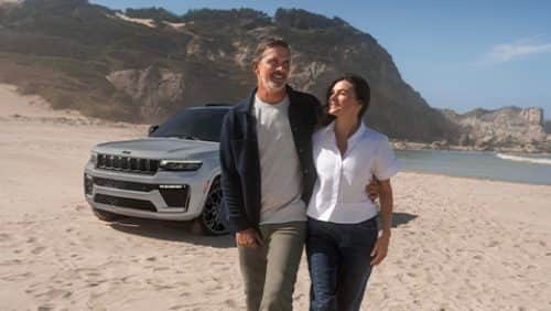 A man and woman walking away from a gray 2027 Jeep Grand Cherokee Summit Obsidian, parked on a sandy beach.