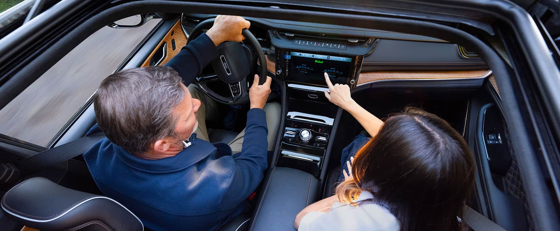 The front seat area of the 2027 Jeep Grand Cherokee Summit, with a man in the driver seat and a woman in the passenger seat. Shot from outside the open sunroof.