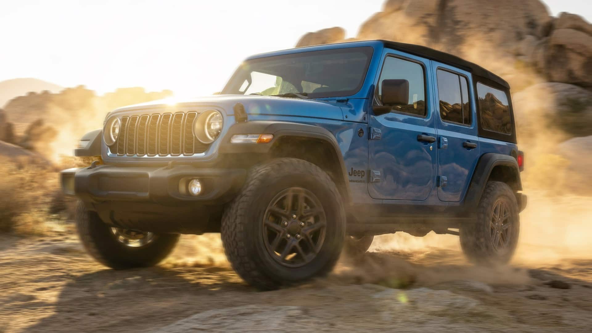 Blue 2027 Jeep Wrangler 4-Door driving through desert dust, showing the seven-slot grille, round headlights and black wheels. Manufacturer photo.