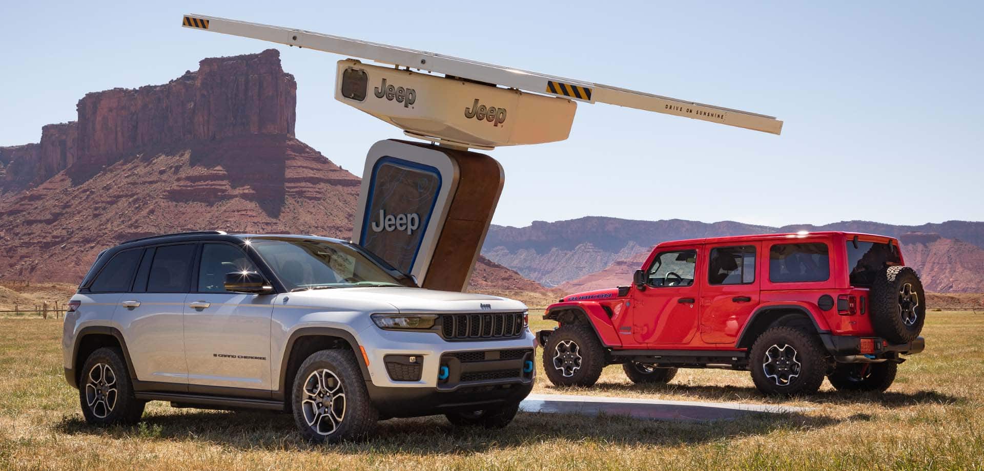 A 2022 Jeep Grand Cherokee Trailhawk 4xe and a 2022 Jeep Wrangler Rubicon 4xe parked at a Jeep-branded charging station in the wilderness.