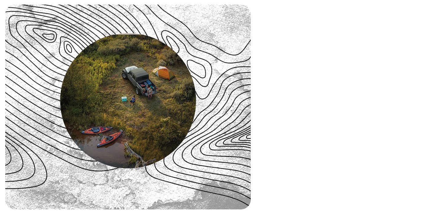 A 2026 Jeep Gladiator Sport S parked beside a lake with camping gear loaded in its pickup box, a man sitting on its open tailgate, a cooler and tent nearby and a woman walking toward the lake where two kayaks are sitting on the edge of the shore.