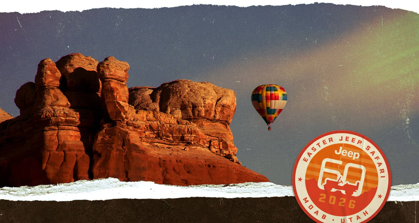A huge rock formation in the desert with snow on the ground and a hot air balloon in the distance. Easter Jeep Safari. Jeep 60 2026. Moab, Utah.