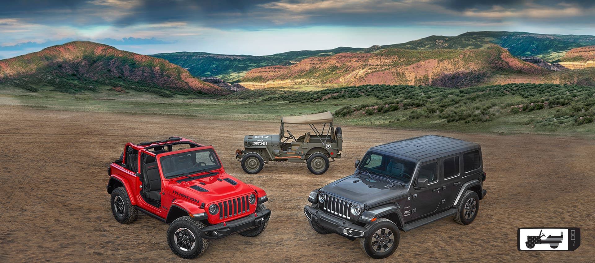 A red 2021 Jeep Wrangler Rubicon, a gray 2021 Wrangler Unlimited Sahara and a 1944 Willys MB parked in a clearing with grassy hills in the background. The Jeep 80th Anniversary logo.