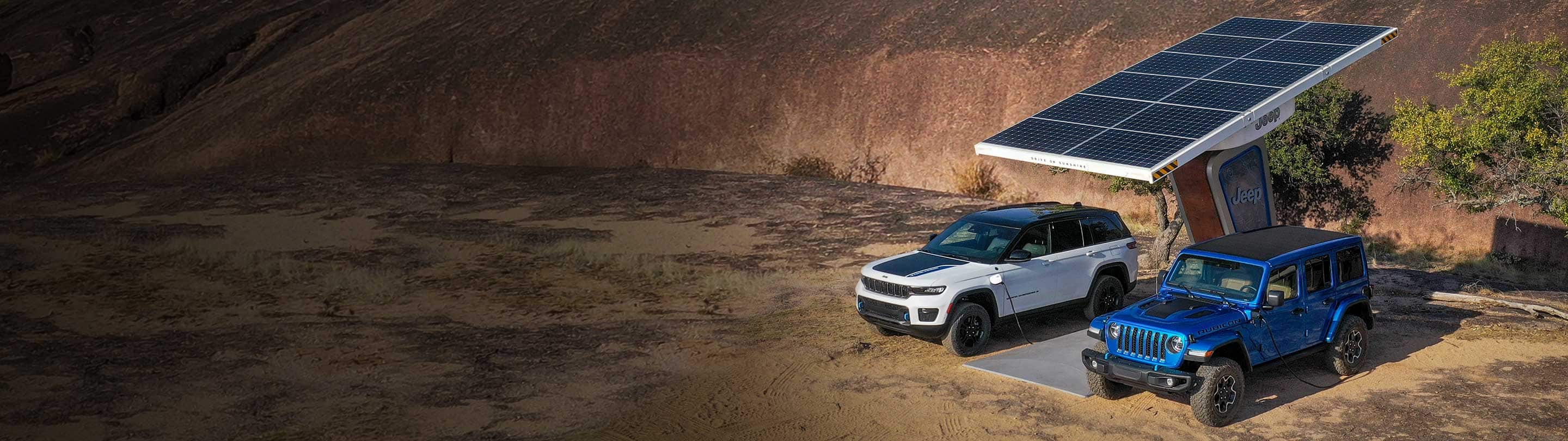 A 2023 Jeep Grand Cherokee Trailhawk 4xe and a 2023 Jeep Wrangler Rubicon 4xe being charged at a Jeep-branded solar-powered charging station located off-road, with a huge sand dune in the background.