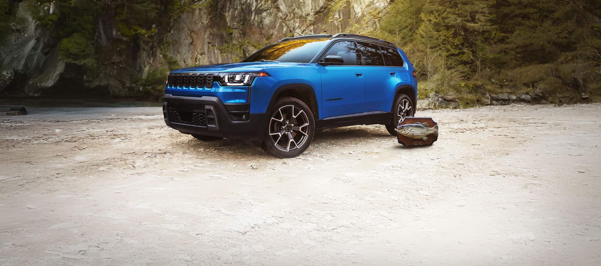 A driver-side front angle of a blue 2026 Jeep Cherokee Overland parked on a clearing with a Billy Bass wall mount beside it and a wooded swamp in the background.
