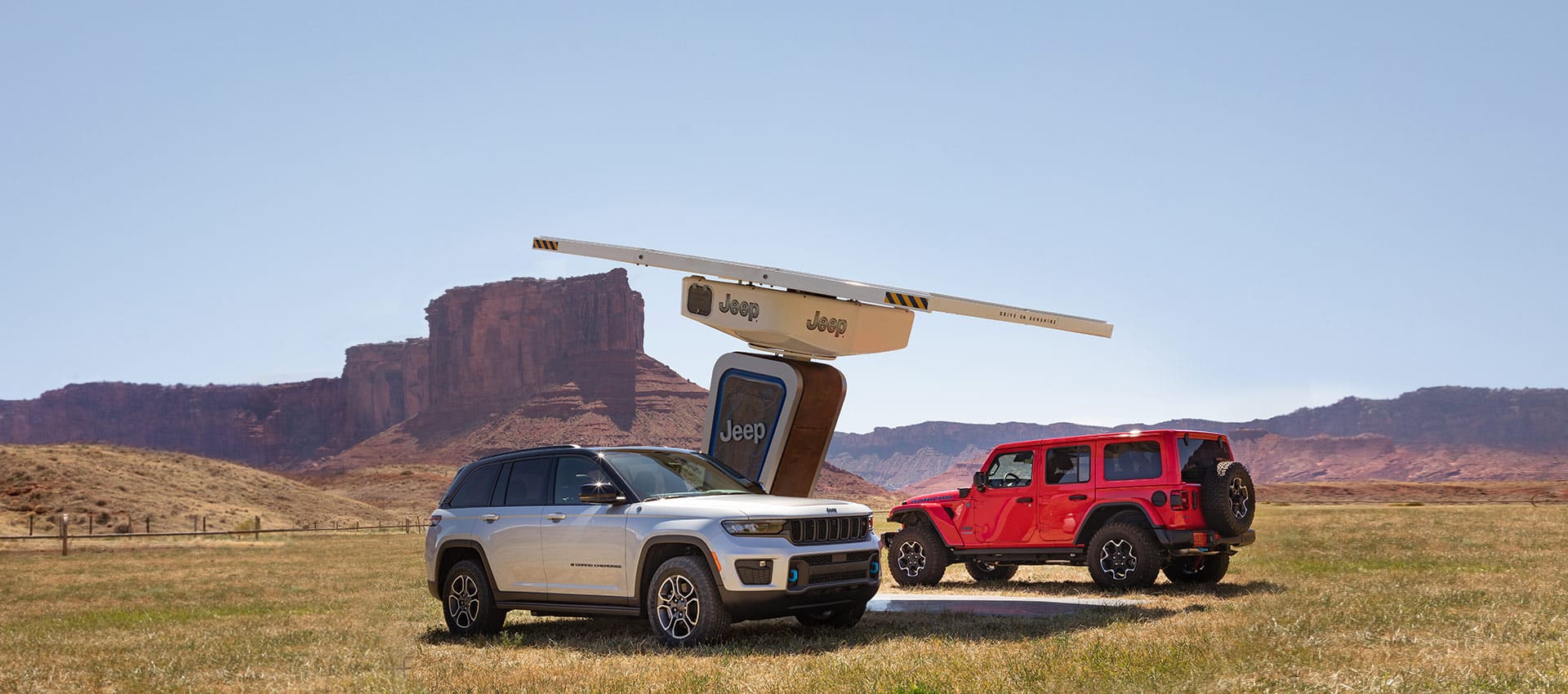 A 2022 Jeep Grand Cherokee Trailhawk 4xe and a 2022 Jeep Wrangler Rubicon 4xe parked off-road at a Jeep-branded solar-powered charging station in the mountains.