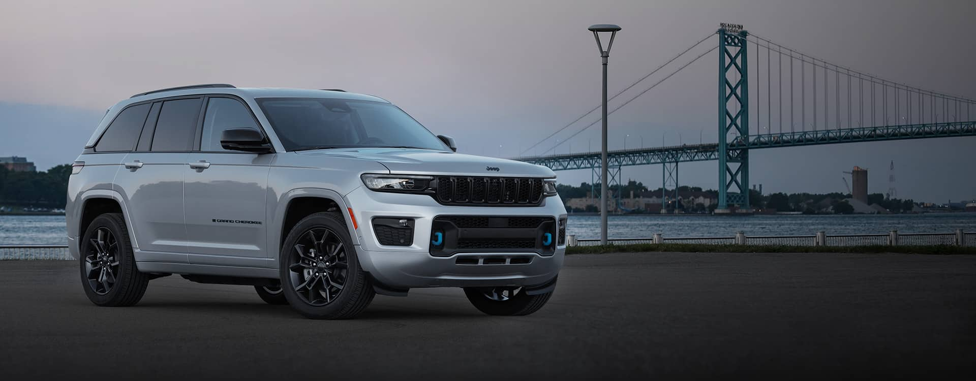 The 2023 Jeep Grand Cherokee 30th Anniversary Edition 4xe parked beside a waterway in the city at twilight with a suspension bridge in the background.