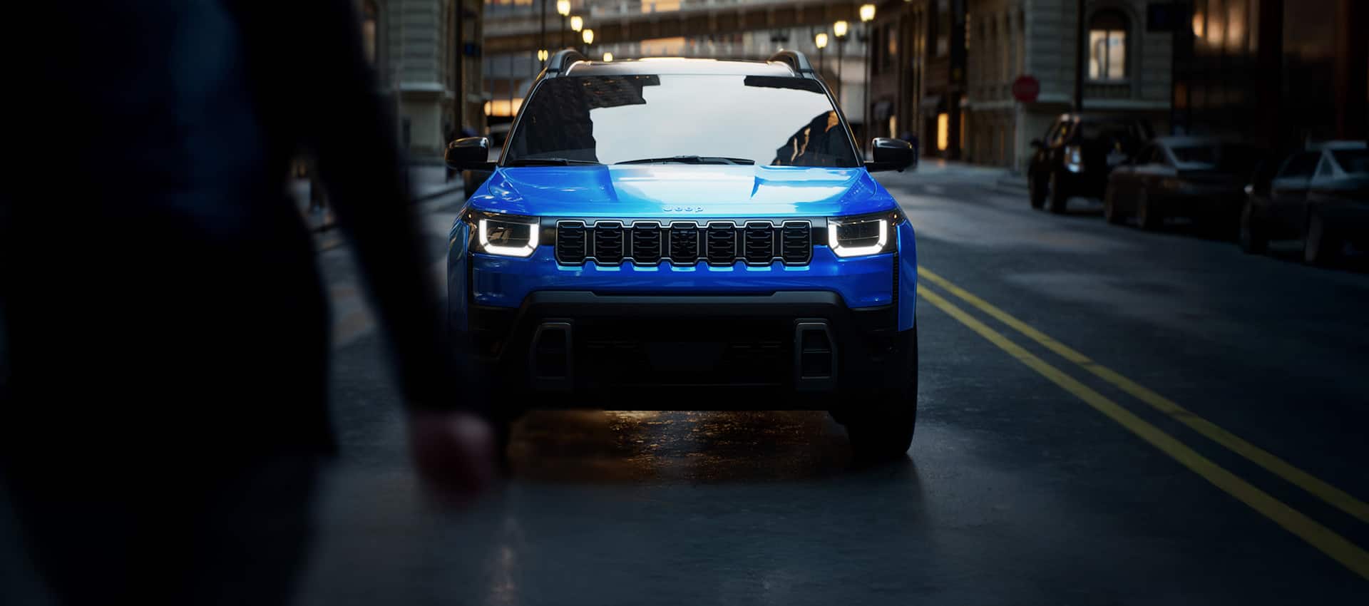 A head-on angle of a blue 2026 Jeep Cherokee Limited traveling on a city street at night.