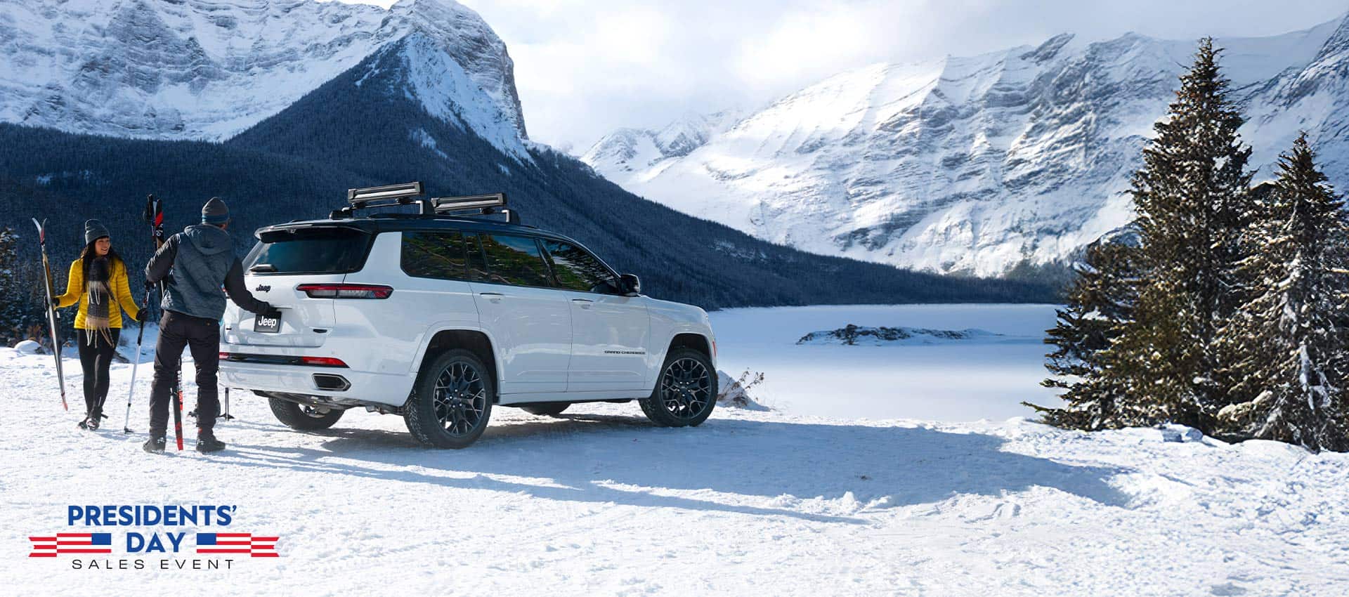 A white 2026 Jeep Grand Cherokee L High Altitude parked on a snow-covered clearing in the mountains. A couple nearby is carrying skiing equipment, and the man is about to open the liftgate. The Presidents' Day Sales Event.