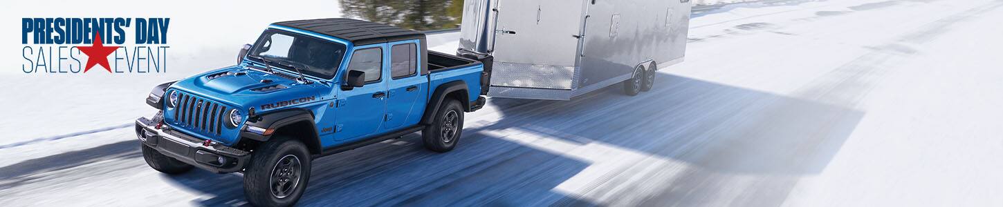 A blue 2023 Jeep Gladiator Rubicon with a black roof, being driven down a snow-covered highway in the mountains, as it tows an enclosed trailer. Jeep. Presidents' Day Sales Event logo.