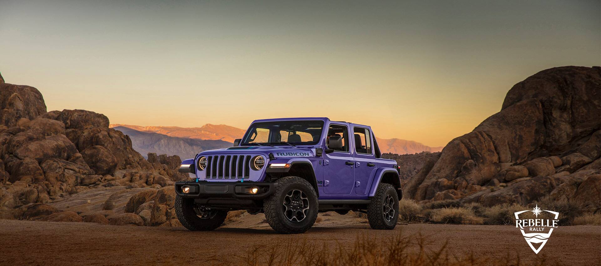 A 2023 Jeep Wrangler Rubicon 4xe parked on a clearing off-road beside rocky hills with mountains in the background at sunset. Rebelle Rally logo.