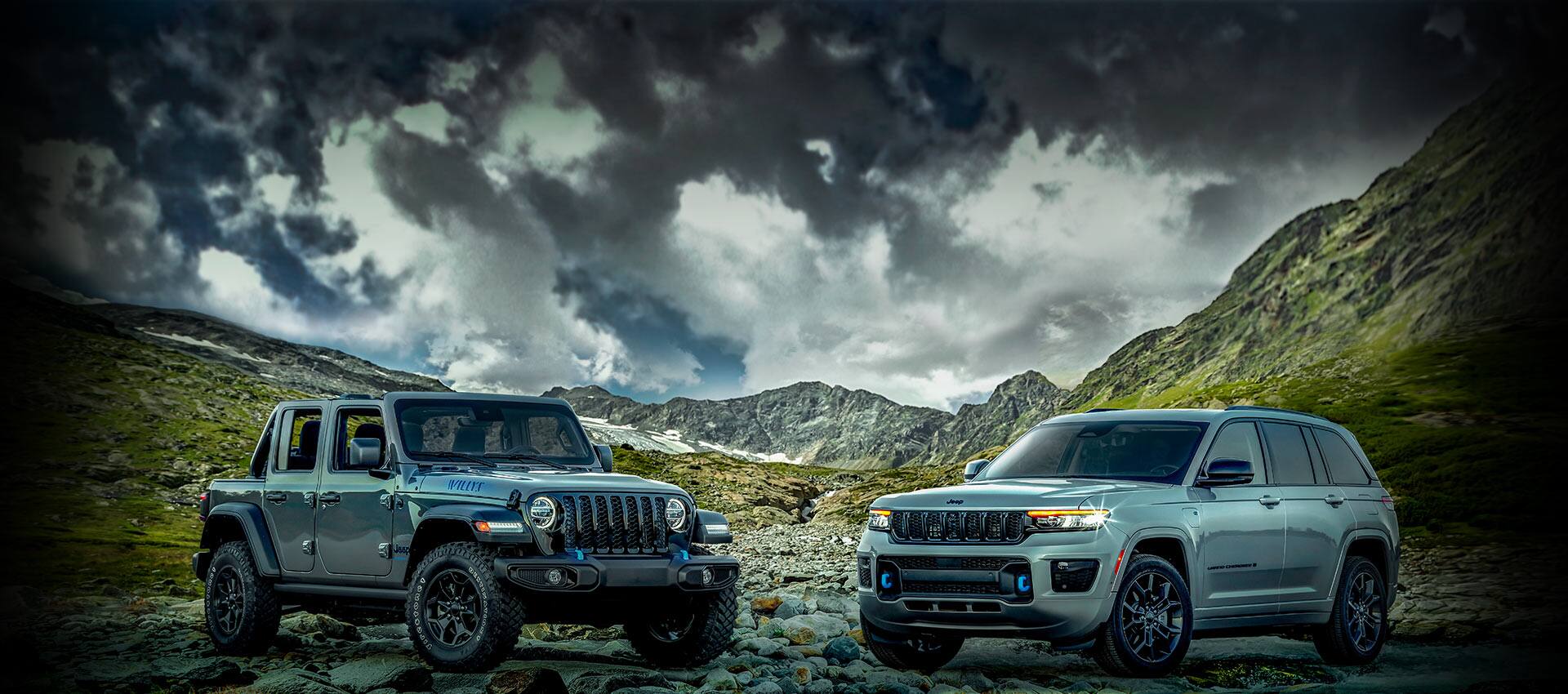 A 2023 Jeep Wrangler Willys 4xe and 2023 Jeep Grand Cherokee 30th Anniversary Edition 4xe parked on a rocky ridge with mountains and a stormy sky in the background.