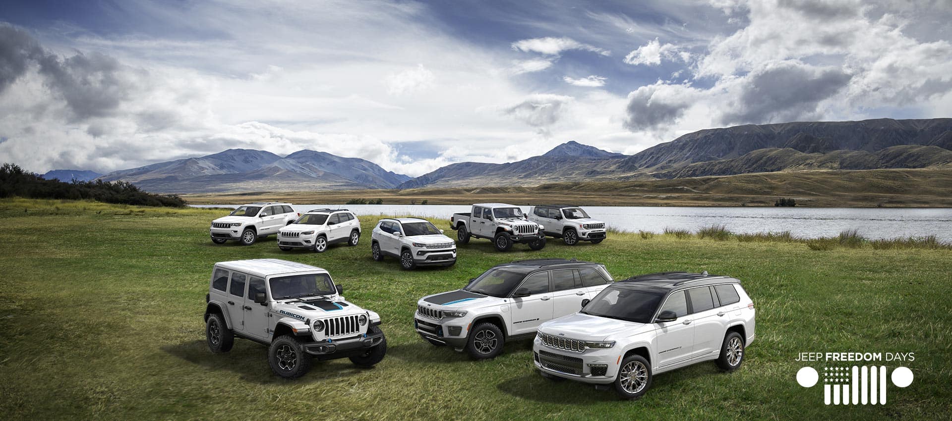 The 2022 Jeep Brand lineup parked beside a lake with mountains in the background. Front row: the Wrangler Unlimited Rubicon 4xe, Grand Cherokee 4xe Trailhawk and Grand Cherokee L. Back row: Grand Cherokee WK Limited, Cherokee Limited, Compass Limited, Gladiator Rubicon and Renegade Limited. The Jeep Freedom Days Logo.