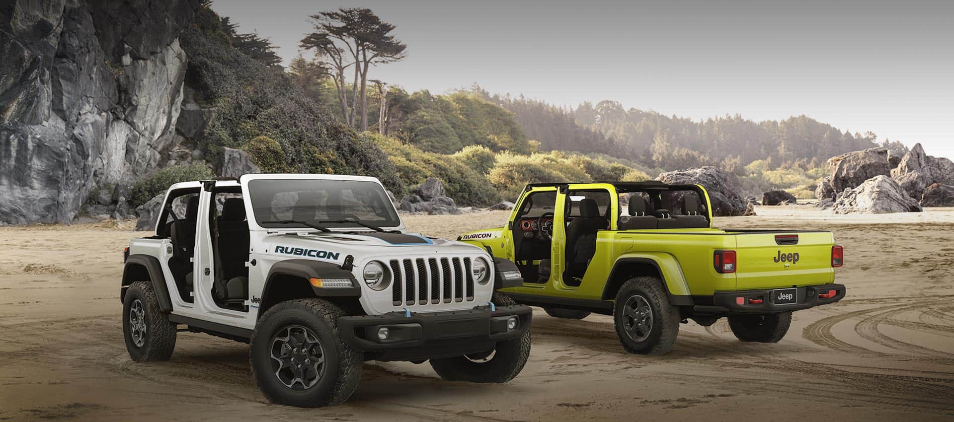 A white 2023 Jeep Wrangler Rubicon 4xe and a lime green 2023 Jeep Gladiator Rubicon, both with their doors and roofs removed, parked opposite of each other on sand at the foot of a large, rocky hill.