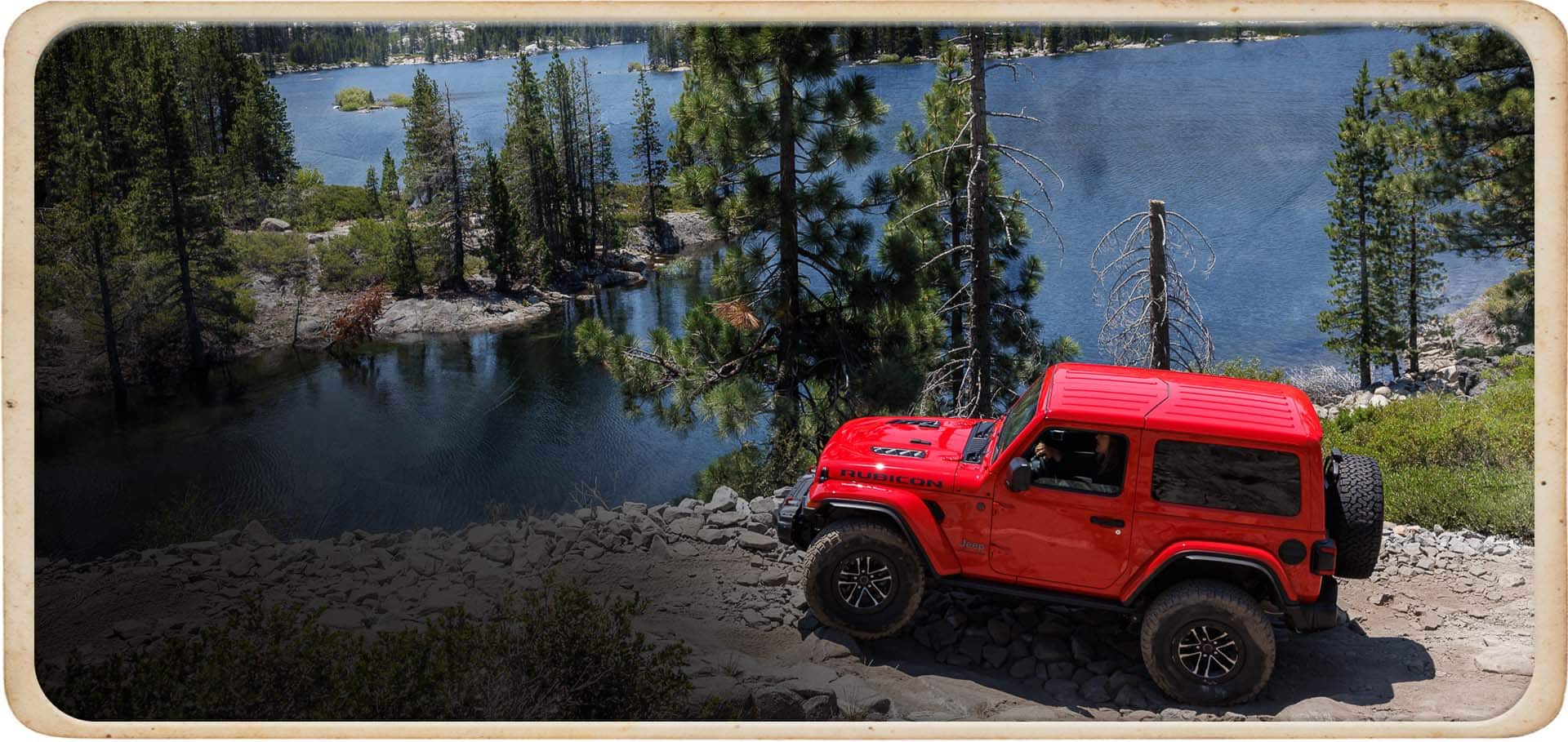 A red 2025 Jeep Wrangler Rubicon X parked on a rocky cliff overlooking Lake Tahoe and a dense forest.