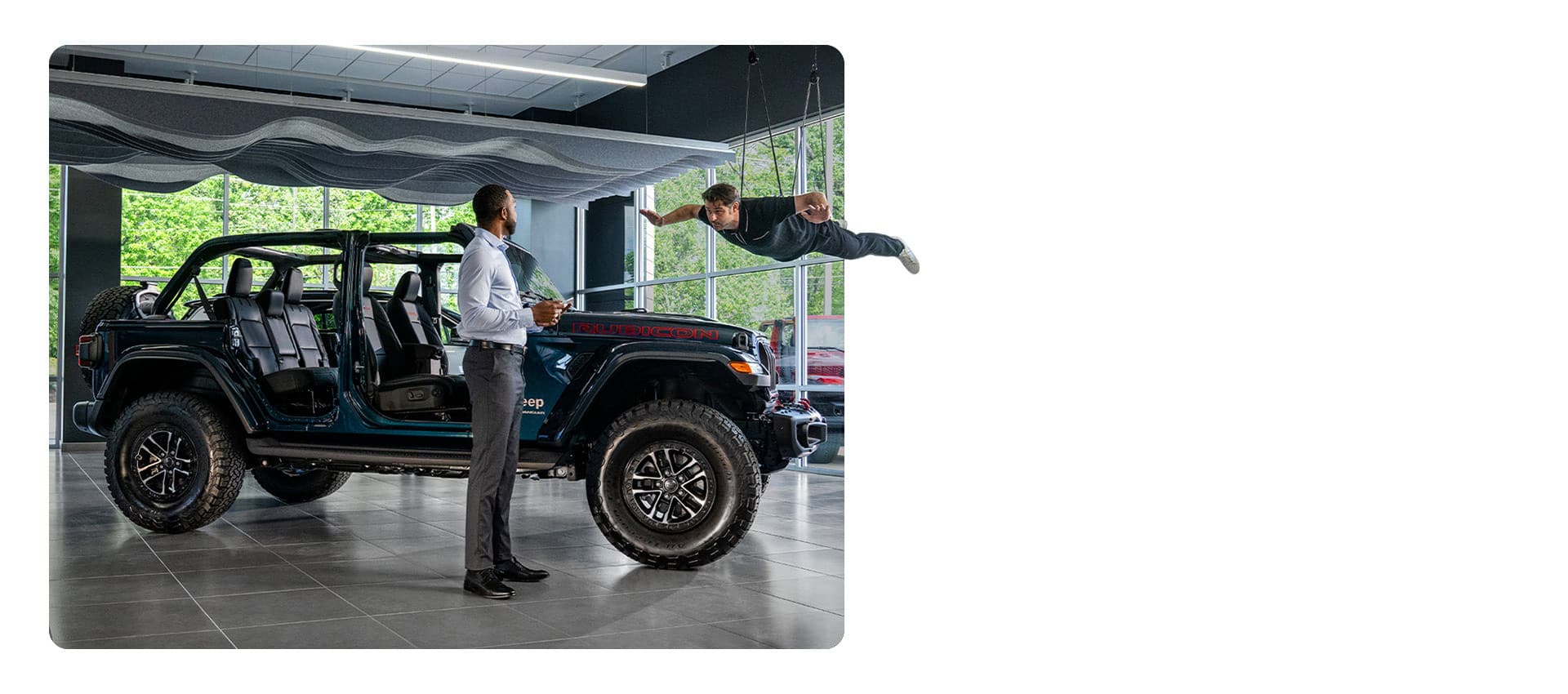 A blue 2025 Jeep Wrangler Rubicon X with its doors and roof removed, on display in a dealer showroom with one man standing nearby and another dangling from wires, as if floating overhead.