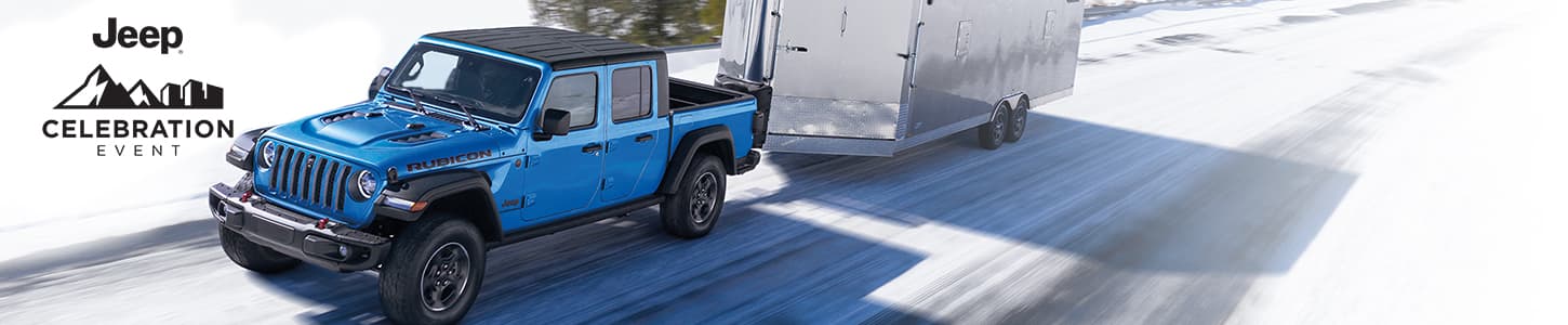 A blue 2023 Jeep Gladiator Rubicon with a black roof, being driven down a snow-covered highway in the mountains, as it tows an enclosed trailer. Jeep. Jeep Celebration Event logo.