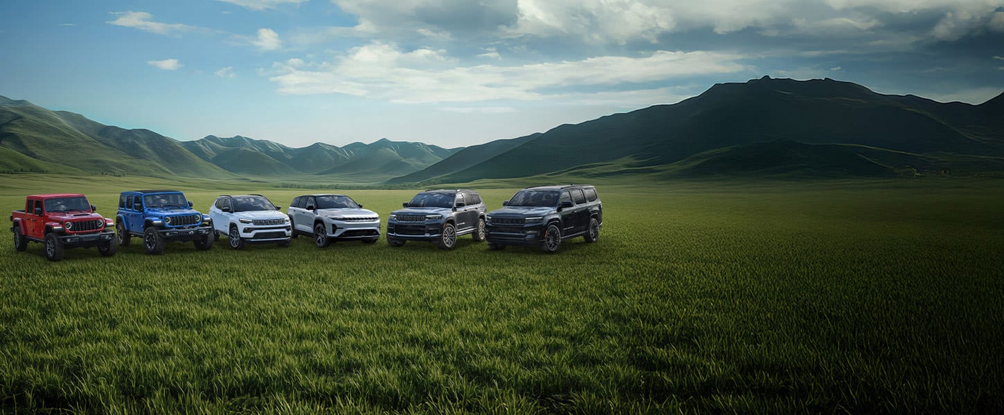 A lineup of six 2025 Jeep Brand vehicles parked on a grassy clearing with mountains in the background. From left to right: a red Gladiator Mojave X, a blue Wrangler Rubicon X 4xe, a white Compass Limited, a silver Wagoneer S Launch Edition, a gray Grand Cherokee Summit Reserve and a black Grand Wagoneer Obsidian.