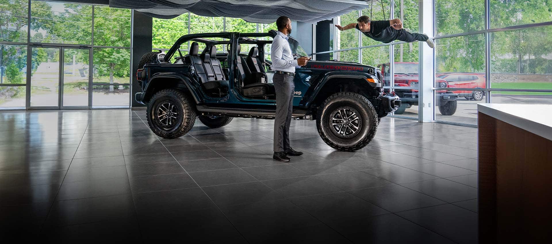 A blue 2025 Jeep Wrangler Rubicon X with its doors and roof removed, on display in a dealer showroom with one man standing nearby and another dangling from wires, as if floating overhead.
