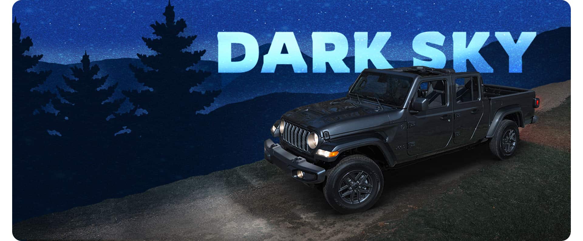 A driver-side front angle of a dark gray 2025 Jeep Gladiator Sport S with the Dark Sky Package on a dirt road with its headlamps on and its roof removed. Dark Sky.