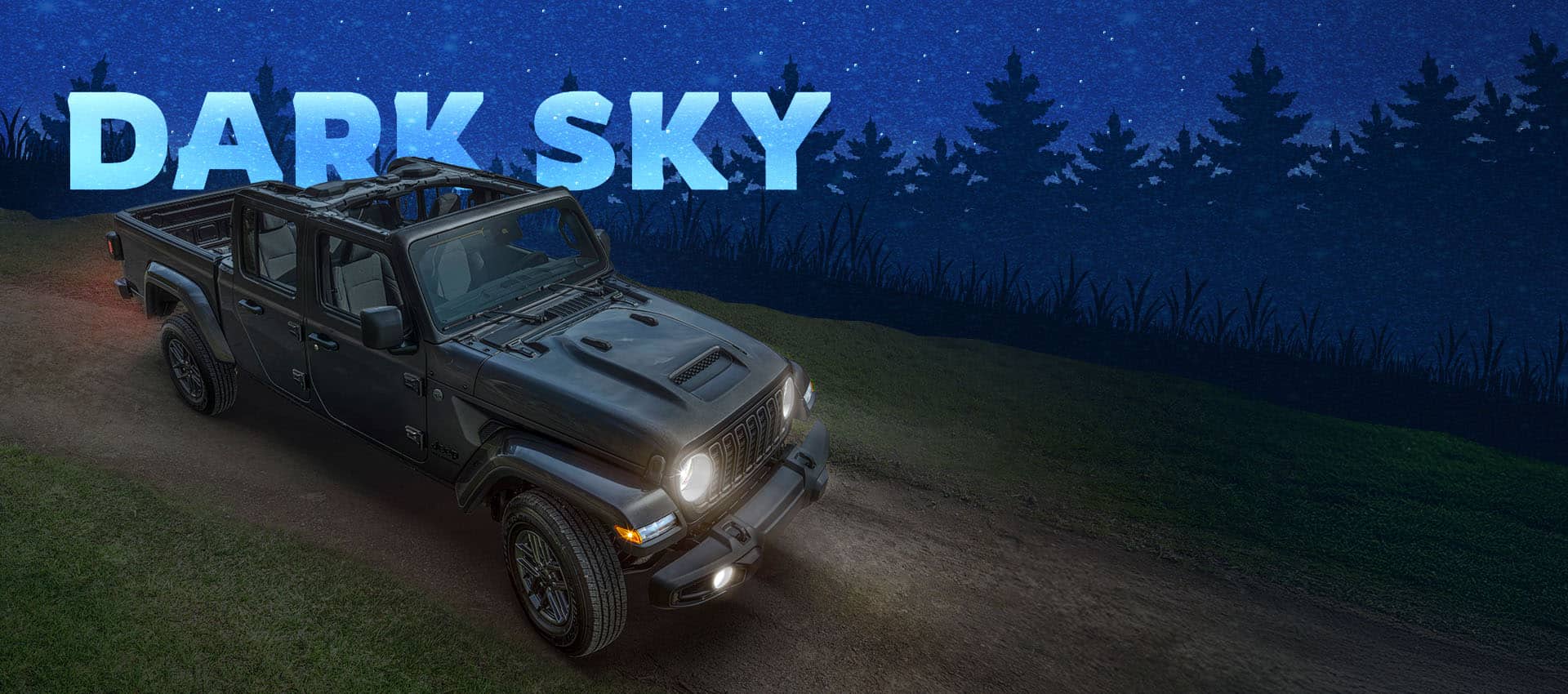 A raised passenger-side front angle of a dark gray 2025 Jeep Gladiator Sport S with the Dark Sky Package on a dirt road with its headlamps on and its roof removed. Dark Sky.