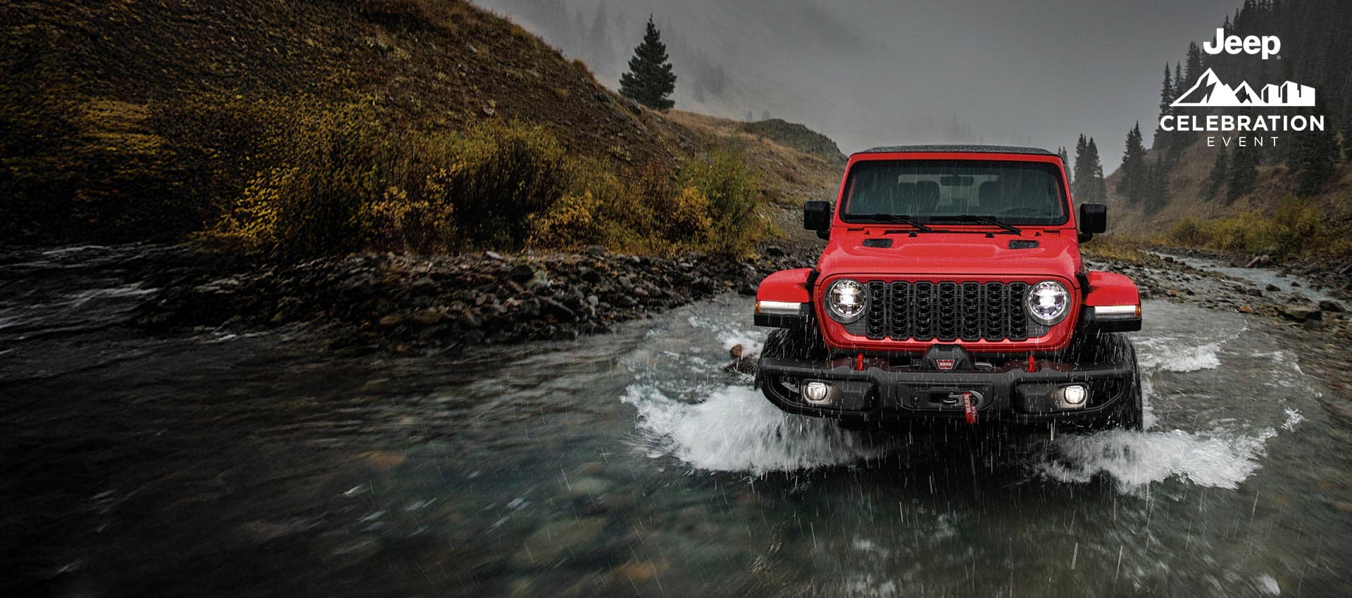 A front angle of a red 2024 Jeep Wrangler Rubicon being driven through a stream, following its path. Jeep Celebration Event.