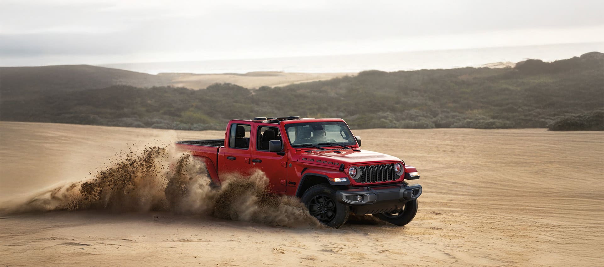 A red 2025 Jeep Gladiator Big Bear kicking up sand as it journeys through the desert, off-road.