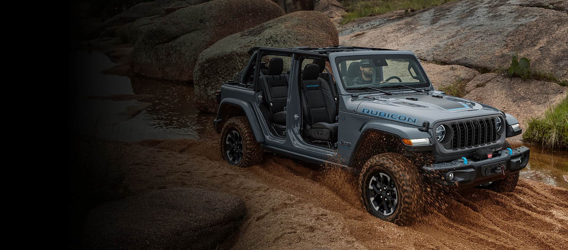 A 2024 Jeep Wrangler Rubicon 4xe with its doors and roof removed, being driven on a sandy trail off-road, with huge boulders on both sides.