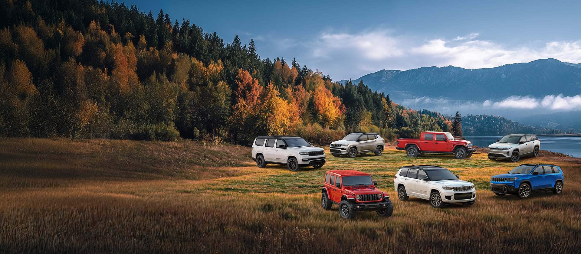 A lineup of six 2025 and one 2026 Jeep Brand vehicles parked on a grassy clearing with a forest, a lake and mountains in the background. In the front row: a red Wrangler Rubicon X, a white Grand Cherokee Summit Reserve and a blue 2026 Cherokee Overland. In the back row: a white Grand Wagoneer Obsidian, a white Compass Limited Altitude, a red Gladiator Mojave X and a white Wagoneer S Launch Edition.