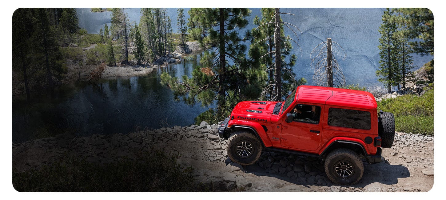A red 2025 Jeep Wrangler Rubicon X parked on a rocky cliff overlooking Lake Tahoe and a dense forest.