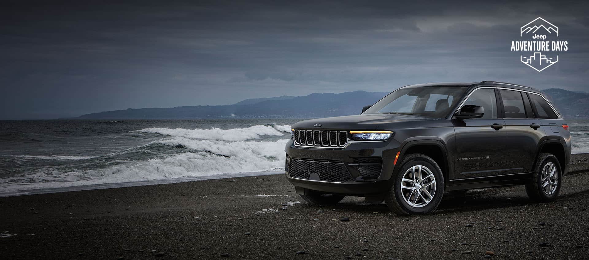An angled driver-side profile of a 2024 Jeep Grand Cherokee Laredo parked on the beach with waves and a storm rolling in. Jeep Adventure Days logo.