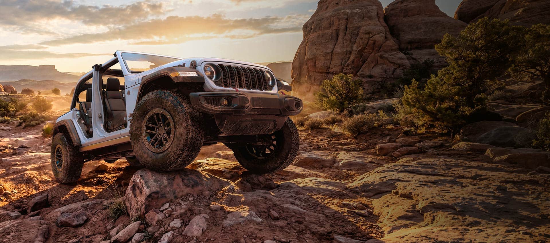 A white 2026 Jeep Wrangler Rubicon with its doors and top removed, crawling over a very rugged trail of bedrock. Drive Into Fall Sales Event.