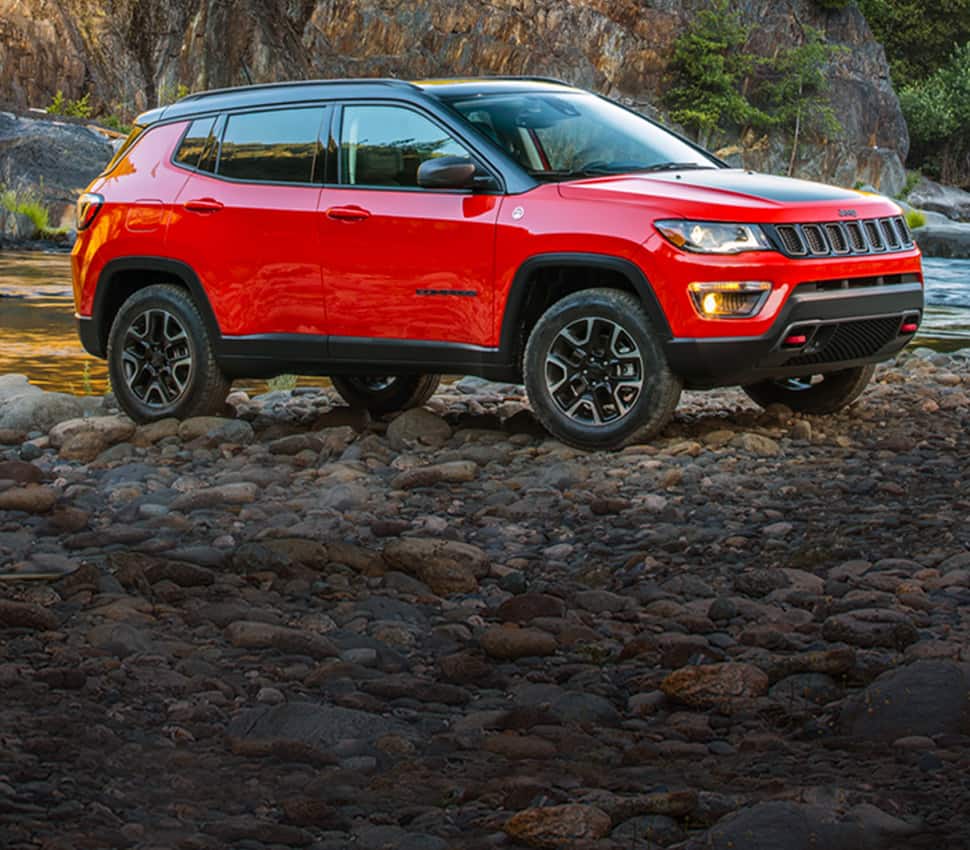 A red 2020 Jeep Compass Trailhawk parked off-road on rocks near a stream.