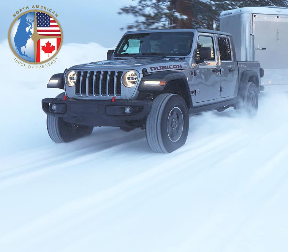 A gray 2020 Jeep Gladiator Rubicon being driven on a snow-covered road, towing a camper.
