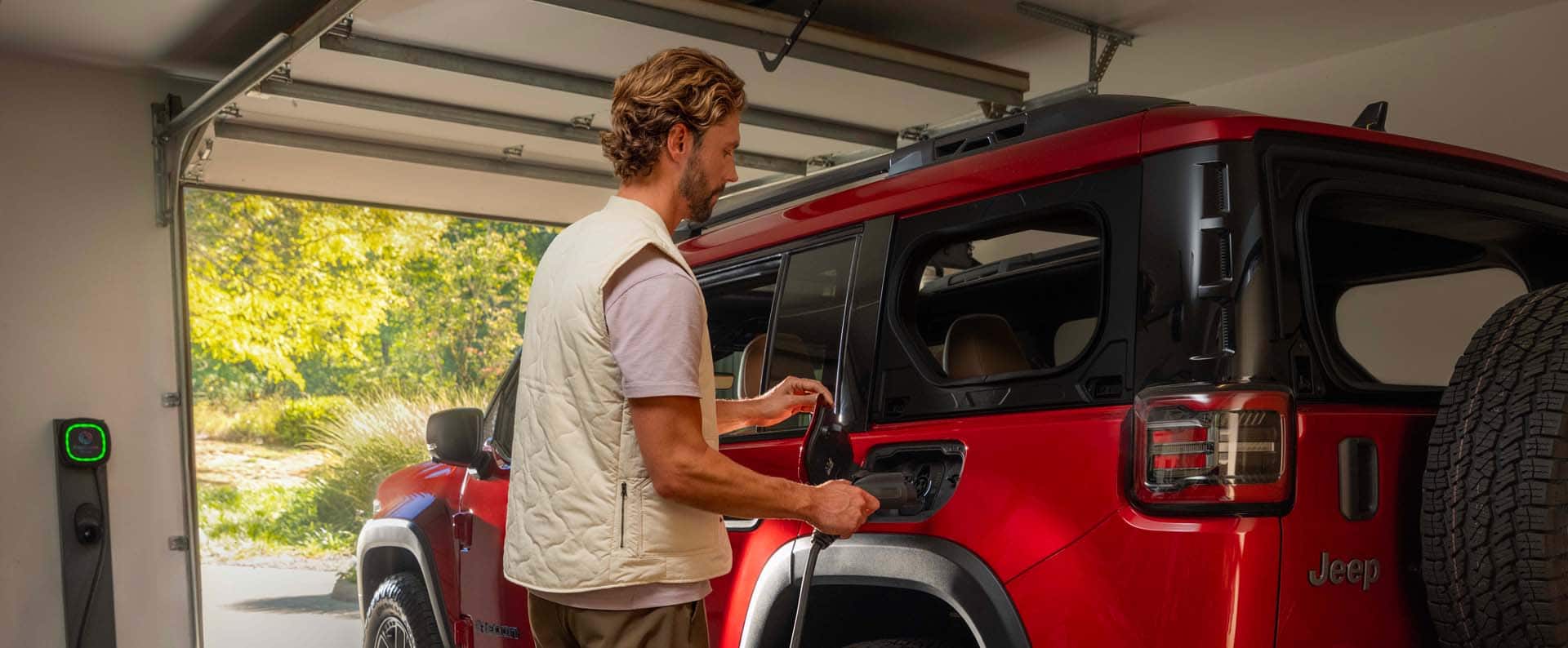A man plugging a Level 2 charging cord into the charging port on a red 2026 Jeep Recon Moab parked in a garage.