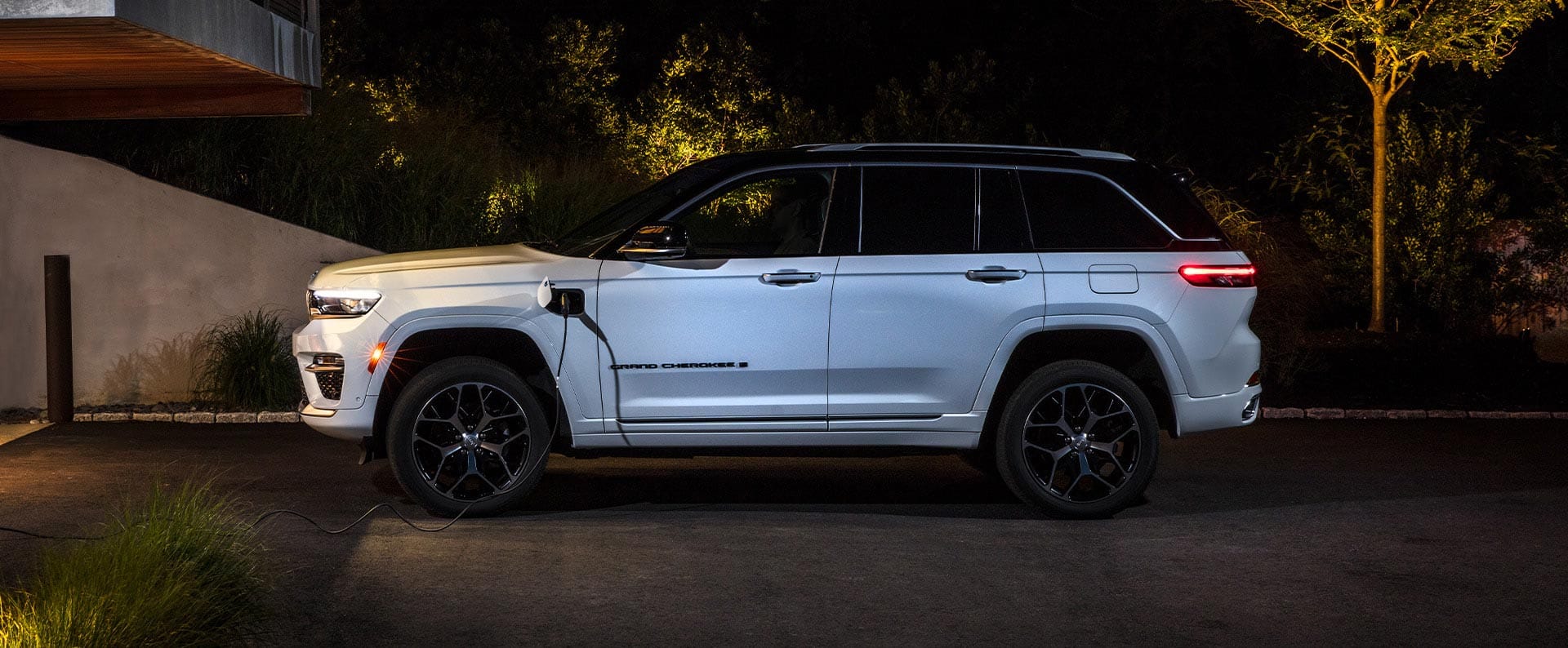 A driver-side profile of a white 2025 Jeep Grand Cherokee 4xe Summit Reserve with a black roof, parked in the driveway of a home, with a charging cord plugged into the vehicle's charging port.