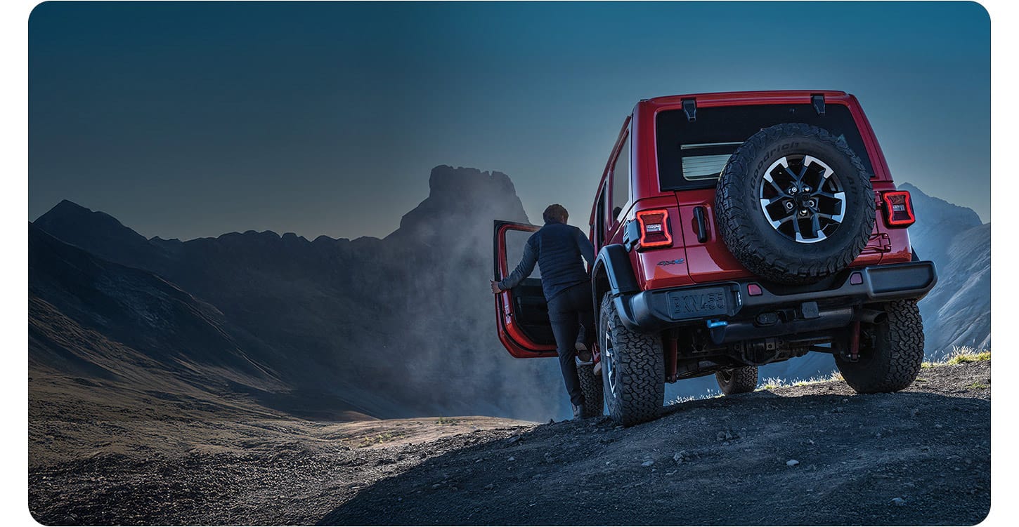 A rear angle of a red 2025 Jeep Wrangler Rubicon 4xe on a cliff, with a man entering opening the driver's door.
