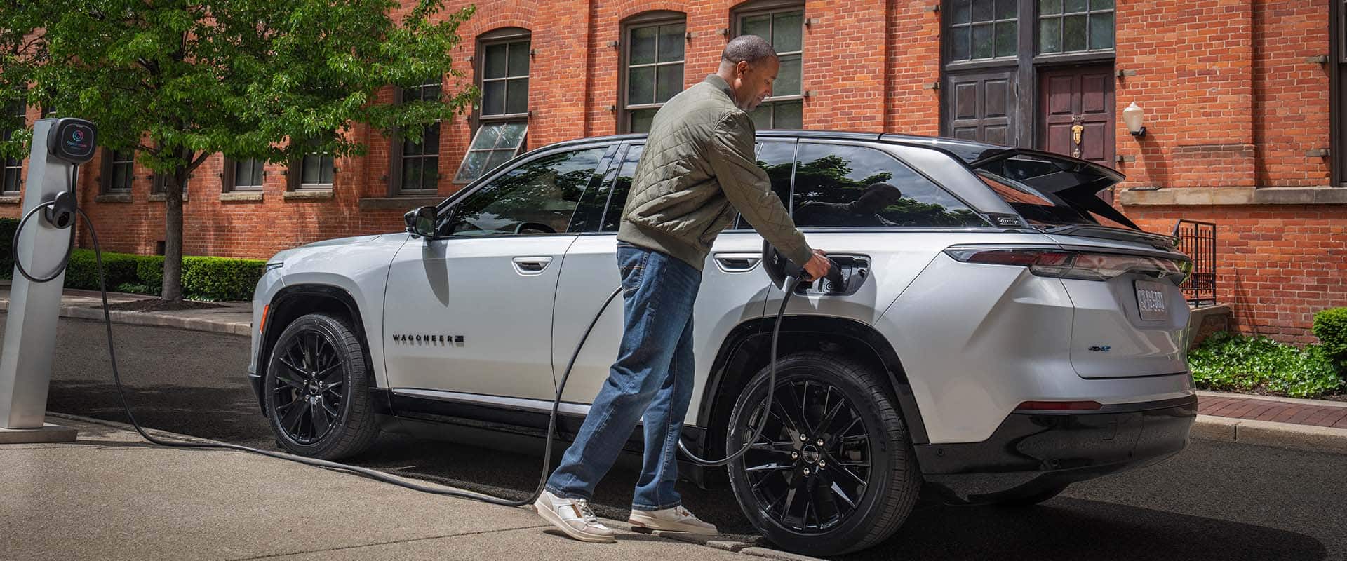 Un Jeep Wagoneer S Launch Edition 2024 plateado estacionado al lado de un edificio de ladrillo, con un hombre enchufando el cable de carga de una estación de carga pública en el puerto de carga del vehículo.