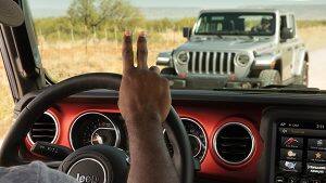 A driver behind the wheel of a Jeep Wrangler performing the Jeep Wave by raising his index and middle finger toward the driver of an approaching Wrangler.