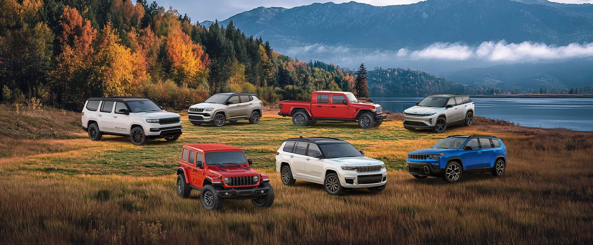 A lineup of six 2025 and one 2026 Jeep Brand vehicles parked on a grassy clearing with a forest, a lake and mountains in the background. In the front row: a red Wrangler Rubicon X, a white Grand Cherokee Summit Reserve and a blue 2026 Cherokee Overland. In the back row: a white Grand Wagoneer Obsidian, a white Compass Limited Altitude, a red Gladiator Mojave X and a white Wagoneer S Launch Edition.