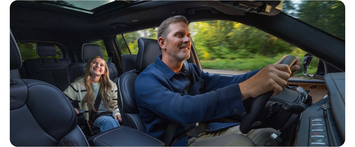 An interior shot of a smiling father driving down a country road in a 3-row Grand Cherokee. His smiling daughter sits in one of the second-row seats.