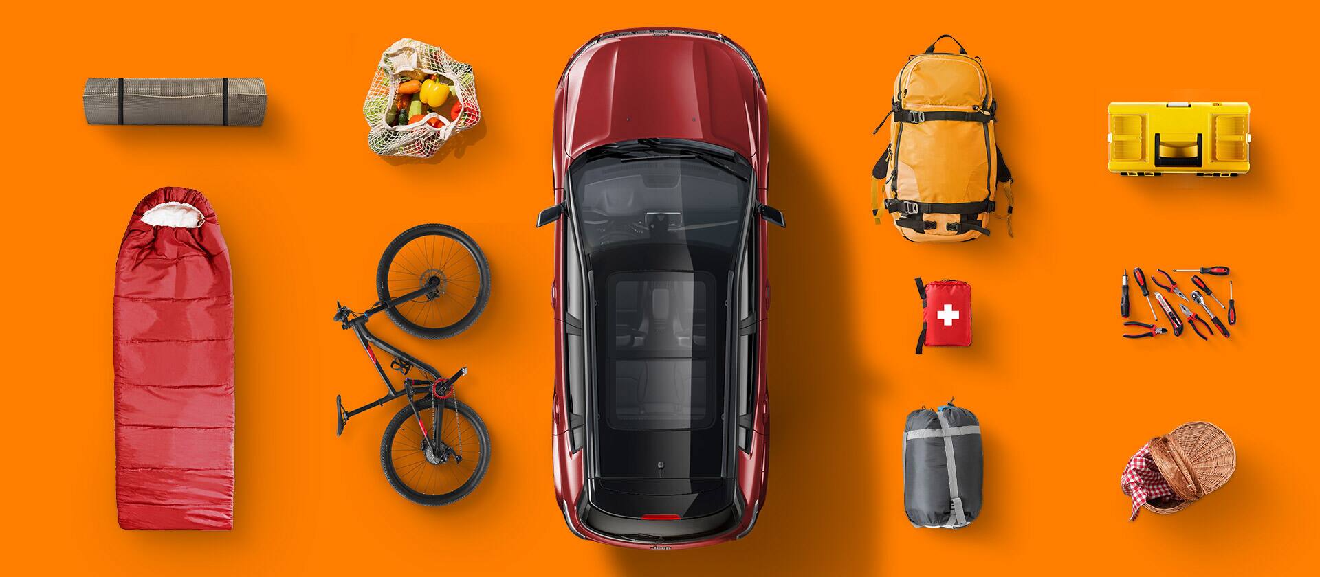 An overhead angle of a red 2026 Jeep Compass Limited displaying the front and rear seats and  cargo area. Outside the vehicle are different types of cargo including a rolled-up rug, a bag of groceries, a sleeping bag, a bicycle, a backpack, tool kit, first aid kit, a set of tools, a duffle and a picnic basket.